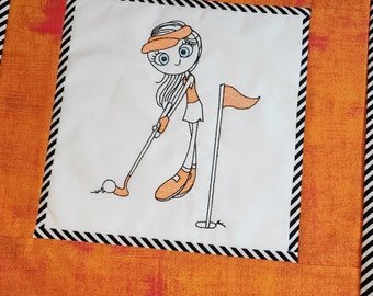 May include: Embroidered design of a girl playing golf. She is wearing a hat, a short-sleeved shirt, and a skirt. She is holding a golf club and is about to hit a golf ball. There is a golf flag in the background.