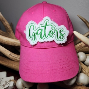 May include: A pink baseball cap with a white and green embroidered patch that says "Gators".