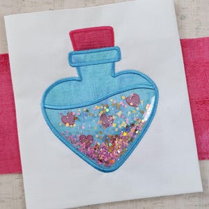 Puede incluir: Una botella azul en forma de corazón con una tapa roja llena de purpurina rosa y dorada y pequeños corazones rosas.
