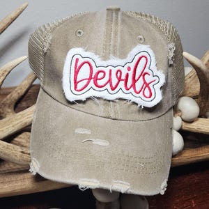 May include: A distressed tan trucker hat with a mesh back. The hat features a white patch with the word "Devils" embroidered in red script with a black outline. The hat has a pre-worn look with distressed details.