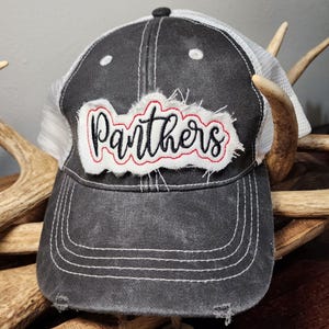 Può includere: Un cappello da camionista nero e bianco con una toppa bianca usurata che dice "Panthers" in lettere corsive nere. La toppa ha cuciture rosse attorno ai bordi.