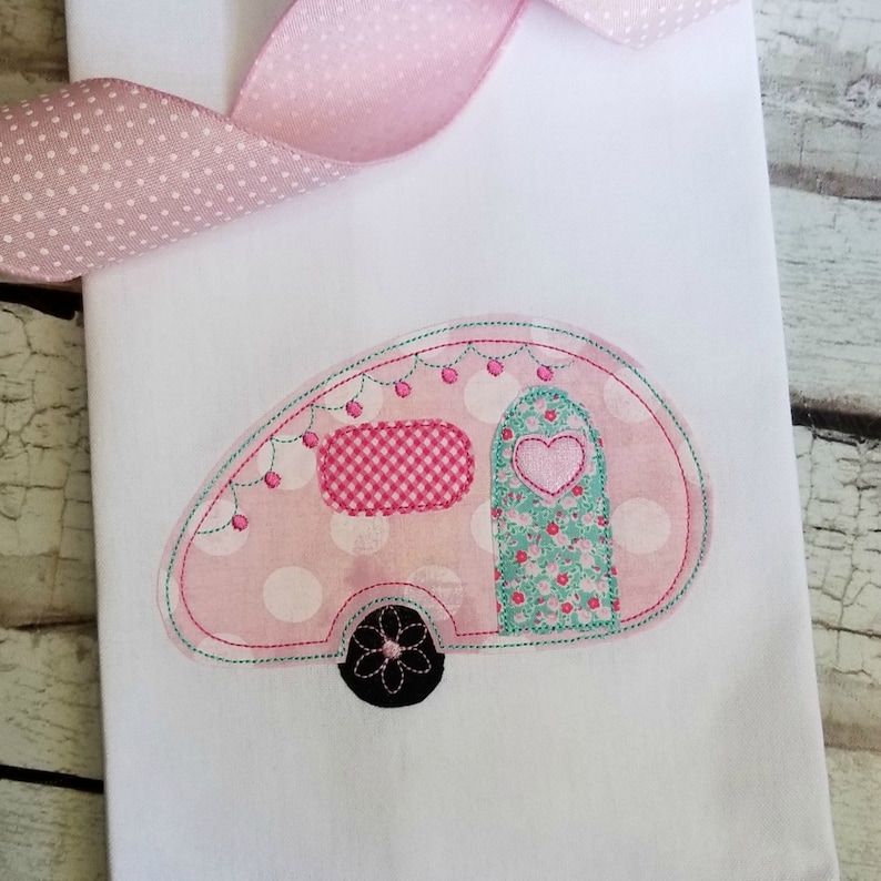 Op de afbeelding: Roze en witte camper met witte stippen, een groene en witte bloemen deur en een roze en wit geruit raam. De camper heeft een zwart wiel met een witte bloem.