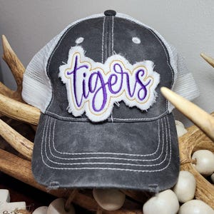 Op de afbeelding: Een zwarte en witte trucker-pet met een verweerde look. De pet heeft een wit mesh-achterpaneel en een paarse geborduurde patch met de tekst "tigers".