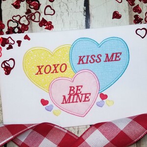 May include: Three heart-shaped candy conversation hearts with the text "XOXO", "Kiss Me", and "Be Mine". The hearts are yellow, blue, and pink.