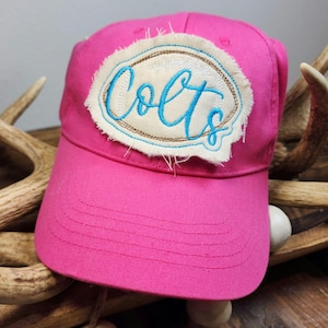 May include: A bright pink baseball cap with the word "Colts" embroidered in light blue script on a cream-colored patch. The patch has a brown border and a distressed, frayed edge.