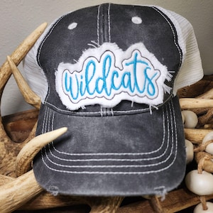 May include: A distressed charcoal gray trucker hat with a white mesh back. The front patch features the word "wildcats" in blue script lettering on a white background with frayed edges. The hat has a curved brim.