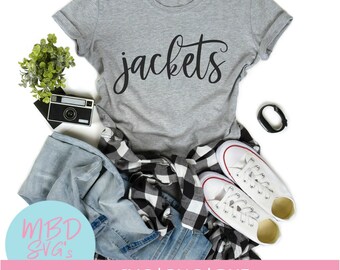 Op de afbeelding: Een grijs T-shirt met het woord "jackets" in zwarte cursieve letters. Het shirt ligt op een witte achtergrond met een paar witte sneakers, een geruit overhemd, een blauwe jeans, een camera en een fitness tracker.