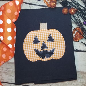 May include: A black kitchen towel with a cheerful orange and white gingham pumpkin face appliqué. The pumpkin has a black stitched smile, eyes, and nose.