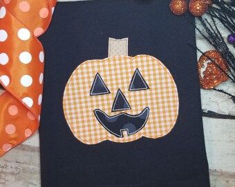 May include: A black kitchen towel with a cheerful orange and white gingham pumpkin face appliqué. The pumpkin has a black stitched smile, eyes, and nose.