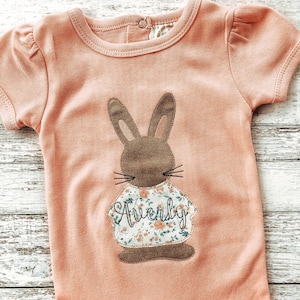 May include: A peach-colored short-sleeved shirt with a brown bunny wearing a floral print shirt with the name "Averly" embroidered on it.