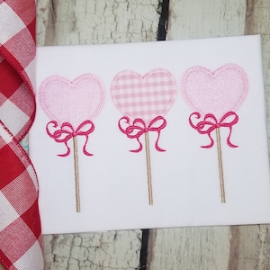 May include: Three pink heart-shaped lollipops with pink bows on sticks. The lollipops are embroidered on a white fabric.