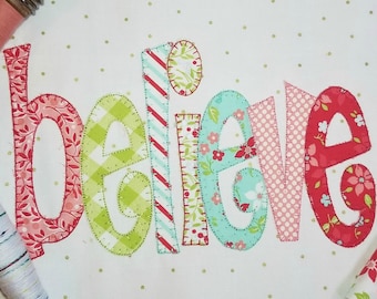 Könnte beinhalten: Eine bunte Stoffapplikation des Wortes „glauben“ in einer verspielten Schriftart. Jeder Buchstabe ist aus einem anderen gemusterten Stoff in Rot-, Grün-, Blau- und Weißtönen gefertigt.