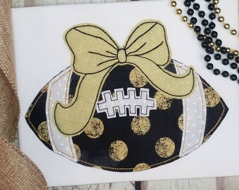 May include: A decorative football design with a gold bow on top. The football is black with gold polka dots and white stripes. A white graphic is in the centre of the football.