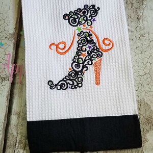 May include: White kitchen towel with a black embroidered high heel shoe design. The shoe is decorated with orange, green, and purple swirls and stars.