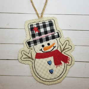 May include: A burlap snowman ornament with a black and white checked hat, a red scarf, and blue hearts. The snowman has a carrot nose and a smiling face.