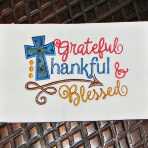 May include: Embroidered design on a white fabric with a blue cross, brown swirls, and the text "Grateful Thankful & Blessed".
