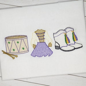 Puede incluir: Diseño bordado de un tambor, un vestido morado y blanco y un par de botas blancas con borlas verdes y doradas. El diseño es perfecto para una banda de marcha o un equipo de baile.