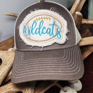 May include: A brown baseball cap with a white mesh back and a curved brim. The cap features a distressed patch with the word "Wildcats" in turquoise script, surrounded by gold and brown stitching.