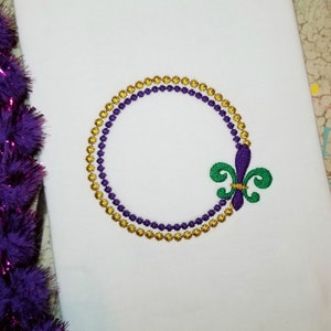 May include: A purple and gold beaded circle frame with a green fleur-de-lis design.