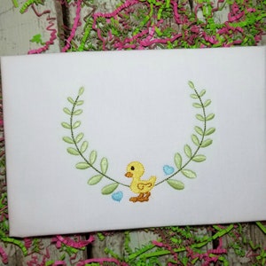 May include: A yellow embroidered chick with blue hearts and a green leafy wreath on a white background.