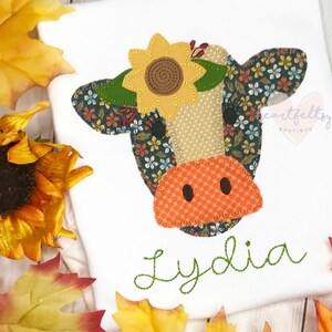 May include: White t-shirt featuring an appliqued cow face with a sunflower and floral pattern. The cow's face is made of various fabrics, including a dotted beige and orange fabric. The name "Lydia" is embroidered in green script.