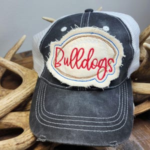 Pode incluir: Boné de caminhoneiro preto e branco desgastado com a palavra "Bulldogs" bordada em vermelho em um patch creme. O patch é contornado com costuras marrons e azuis. O boné tem uma parte traseira de malha e uma aba curva.