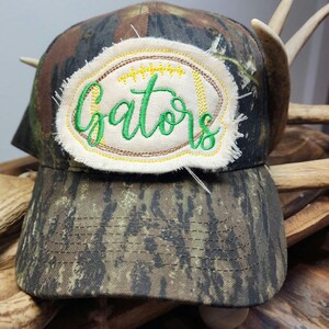 May include: Camouflage baseball cap with a patch that reads "Gators" in green script. The patch is oval-shaped with a frayed edge and yellow and brown stitching. The hat has a curved brim.