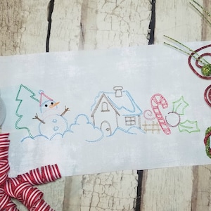 Puede incluir: Diseño bordado con un muñeco de nieve, una casa, un bastón de caramelo y acebo. El diseño está bordado en hilo azul, verde, rojo y marrón sobre una tela blanca.