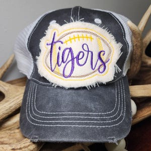Pode incluir: Boné de caminhoneiro cinza e branco com um visual desgastado. O patch frontal apresenta um design de futebol americano com a palavra "tigers" bordada em roxo. A bola tem costuras amarelas.