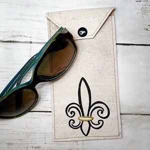 May include: A light-colored cork sunglasses case with a black fleur-de-lis design and a black button closure. The case holds a pair of sunglasses with a black and teal frame.