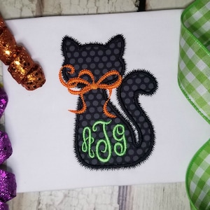 Puede incluir: Gato negro con lazo naranja y monograma verde "JTG" en una camiseta blanca.