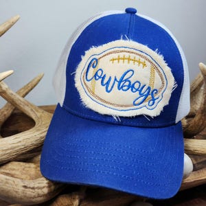Pode incluir: Boné de caminhoneiro azul e branco com a palavra "Cowboys" bordada em um patch em forma de bola de futebol. O boné tem um painel frontal azul e uma parte traseira de malha branca. O patch é creme com costuras marrons e douradas.