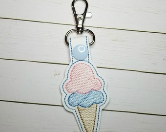 Può includere: Un portachiavi a forma di cono gelato rosa e blu con un moschettone argentato. Il cono gelato è ricamato su un tessuto bianco.