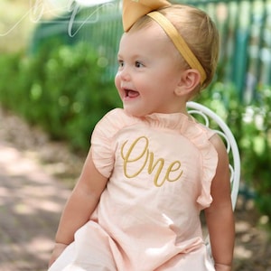 May include: A baby girl wearing a pink dress with a gold embroidered "One" on the front. She is wearing a gold headband and smiling.