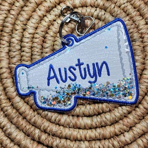 Puede incluir: Un llavero de megáfono de animadora azul y blanco con el nombre "Austyn" en escritura azul. El llavero está decorado con purpurina y tiene un llavero plateado.