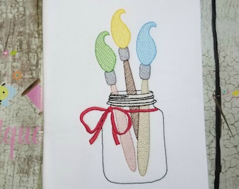 May include: Three paintbrushes in a clear glass jar with a red ribbon tied around the top. The paintbrushes are yellow, green, and blue.