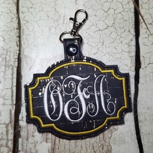 Diseño de bordado con pestaña a presión en el bastidor para monogramas, llaveros y etiquetas de bolsos.