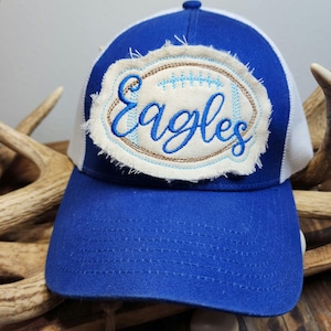 May include: A blue and white baseball cap with the word "Eagles" embroidered on the front. The word is in a cursive font and is surrounded by a football-shaped patch with a distressed edge. The cap has a mesh back.