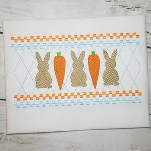 May include: White fabric with embroidered Easter-themed design. Features two bunnies and two carrots in the center, with a repeating pattern of orange and blue stitching. The bunnies are a burlap-like texture.