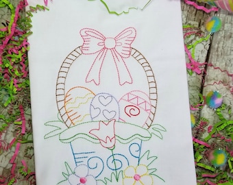 May include: Embroidered design of an Easter basket with a pink bow, filled with colorful eggs and a flower. The basket is surrounded by green leaves and flowers. The word "Egg" is embroidered below the basket.
