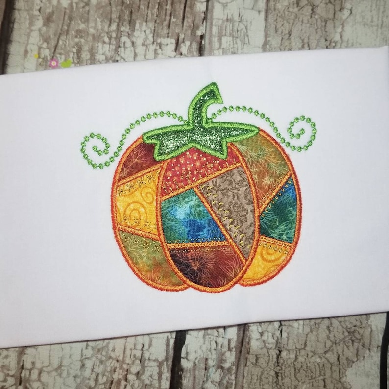K&ouml;nnte beinhalten: Ein Patchwork-K&uuml;rbis mit einem gr&uuml;nen Stiel und Bl&auml;ttern. Der K&uuml;rbis besteht aus verschiedenen Stoffen in den Farben Orange, Gelb, Braun, Blau und Gr&uuml;n. Der K&uuml;rbis ist mit orangefarbenem Garn umrandet.