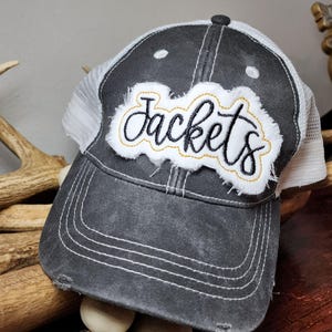 Puede incluir: Una gorra de camionero negra y blanca con una malla blanca en la parte posterior. La gorra tiene un aspecto desgastado y presenta un parche blanco con la palabra "Jackets" bordada en negro.