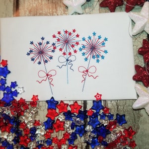 Op de afbeelding: Drie geborduurde vuurwerk in rood, wit en blauw met strikken op een witte achtergrond.