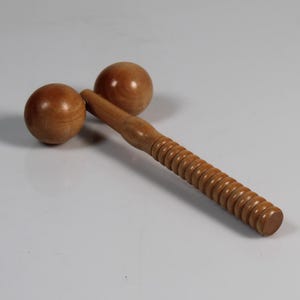 Könnte beinhalten: Ein Massagegerät aus Holz mit zwei kugelförmigen Köpfen und einem geriffelten Griff. Das Werkzeug besteht aus hellbraunem Holz und ist für Muskelentspannung und Therapie konzipiert. Das Werkzeug ist etwa 20 cm lang.