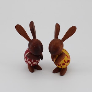 May include: Two wooden bunny figurines with brown bodies and long ears. One bunny wears a red and white patterned outfit, the other a yellow and orange patterned outfit. The bunnies are standing on a white surface.