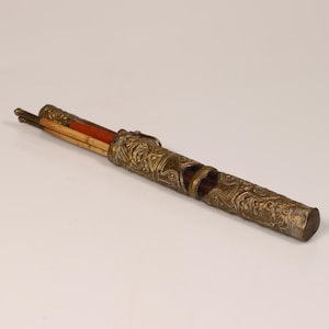 May include: An antique flute in a decorative case. The flute is made of wood and metal, with a reddish-brown body and a silver-colored metal case. The case has intricate floral patterns and a cylindrical shape.