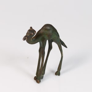 May include: A small, bronze-coloured camel figurine with a slender build. The camel is depicted in a walking pose, with its head slightly lowered. The figurine has a weathered appearance, suggesting an antique or vintage piece.