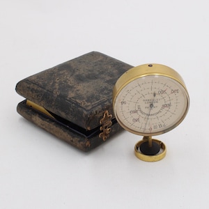 Könnte beinhalten: Ein antikes Messingbarometer mit cremefarbenem Zifferblatt und schwarzen Markierungen, neben einem dunkelbraunen ledergebundenen Etui. Das Barometer steht auf einem kleinen Ständer. Das Etui ist geschlossen.