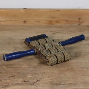 Könnte beinhalten: Ein Massagegerät mit einer Reihe kleiner, strukturierter Rollen und zwei blauen Griffen. Die Rollen sind in einer Reihe angeordnet. Die Griffe sind aus Holz und blau lackiert. Das Werkzeug ist für Massagen konzipiert.