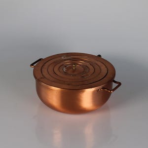 Könnte beinhalten: Ein kleiner, kupferfarbener Kochtopf mit passendem Deckel. Der Deckel hat eine Reihe konzentrischer Kreise und einen kleinen Knopf in der Mitte. Der Topf hat zwei kleine Griffe auf beiden Seiten.